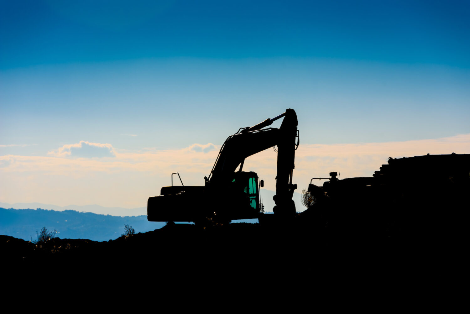 Backlit silhouette of an excavator on top of a hill with unfocused sky background
